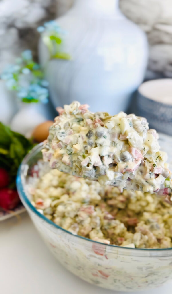 Russian Olivier Salad - Simple Potato Salad in a blue patterend bowl accented with fresh dill with raddish with green leaves and brown eggs in a platter on the side with a blue accented plant in background.