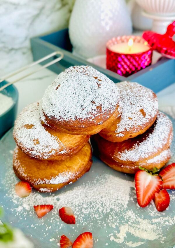 Nutella and Strawberry Filled Donuts (Ponchiks) donuts piled up ontop of eachother as a layer of ponchik donuts sprinkled with powdered surgar over them with fresh strawberry sliced on a blueish grey colored plate with powdered sugar sprinkled around plate. Grey tray in background with a hot pink lit candle with candle lid leaning on the edge of the candle as a accent decor. White canisters along with a white pitcher filled with white flowers with yellow center in flower in pitcher. Grey bowl the the left side of the picture with powdered sugar and powdered sugar sifter resting on powder sugar in bowl with a white towel and floral leaves accenting around photo on a white countertop.