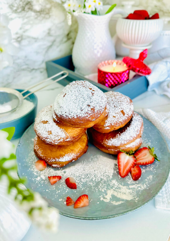 Nutella and Strawberry Filled Donuts (Ponchiks) donuts piled up ontop of eachother as a layer of ponchik donuts sprinkled with powdered surgar over them with fresh strawberry sliced on a blueish grey colored plate with powdered sugar sprinkled around plate. Grey tray in background with a hot pink lit candle with candle lid leaning on the edge of the candle as a accent decor. White canisters along with a white pitcher filled with white flowers with yellow center in flower in pitcher. Grey bowl the the left side of the picture with powdered sugar and powdered sugar sifter resting on powder sugar in bowl with a white towel and floral leaves accenting around photo on a white countertop.