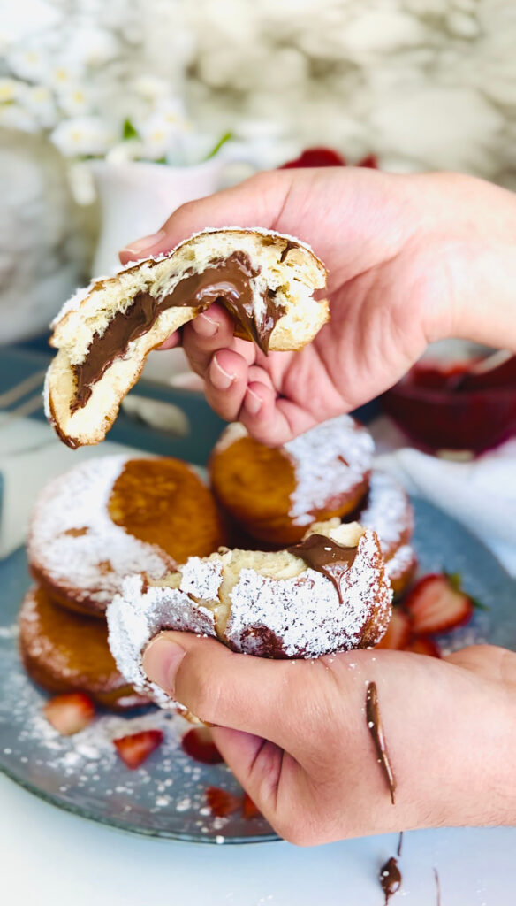Nutella and Strawberry Filled Donuts (Ponchiks) two hands pulling apart ponchik with nutella spilling out of it and spilling on hand. Donuts piled up ontop of eachother as a layer of ponchik donuts sprinkled with powdered surgar over them with fresh strawberry sliced on a blueish grey colored plate with powdered sugar sprinkled around plate. Grey tray in background with a hot pink lit candle with candle lid leaning on the edge of the candle as a accent decor. White canisters along with a white pitcher filled with white flowers with yellow center in flower in pitcher. Grey bowl the the left side of the picture with powdered sugar and powdered sugar sifter resting on powder sugar in bowl with a white towel and floral leaves accenting around photo on a white countertop.