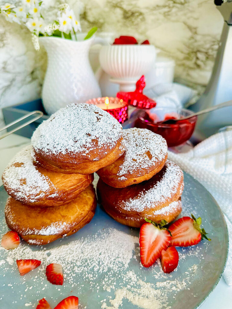 Nutella and Strawberry Filled Donuts (Ponchiks) donuts piled up ontop of eachother as a layer of ponchik donuts sprinkled with powdered surgar over them with fresh strawberry sliced on a blueish grey colored plate with powdered sugar sprinkled around plate. Grey tray in background with a hot pink lit candle with candle lid leaning on the edge of the candle as a accent decor. White canisters along with a white pitcher filled with white flowers with yellow center in flower in pitcher. Grey bowl the the left side of the picture with powdered sugar and powdered sugar sifter resting on powder sugar in bowl with a white towel and floral leaves accenting around photo on a white countertop.