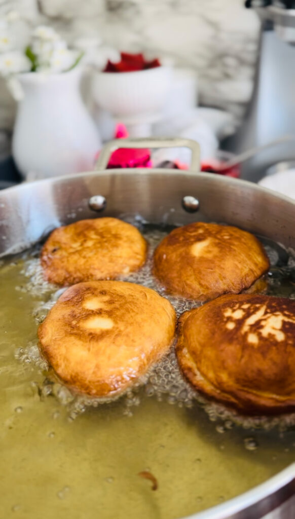 Nutella and Strawberry Filled Donuts (Ponchiks) Frying ponchiks in oil in a frying pan filled with oil. White with yellow center flowers accenting photo with a blue place with sifter laying ontop. Pink candle with white pitcher and bown in background.