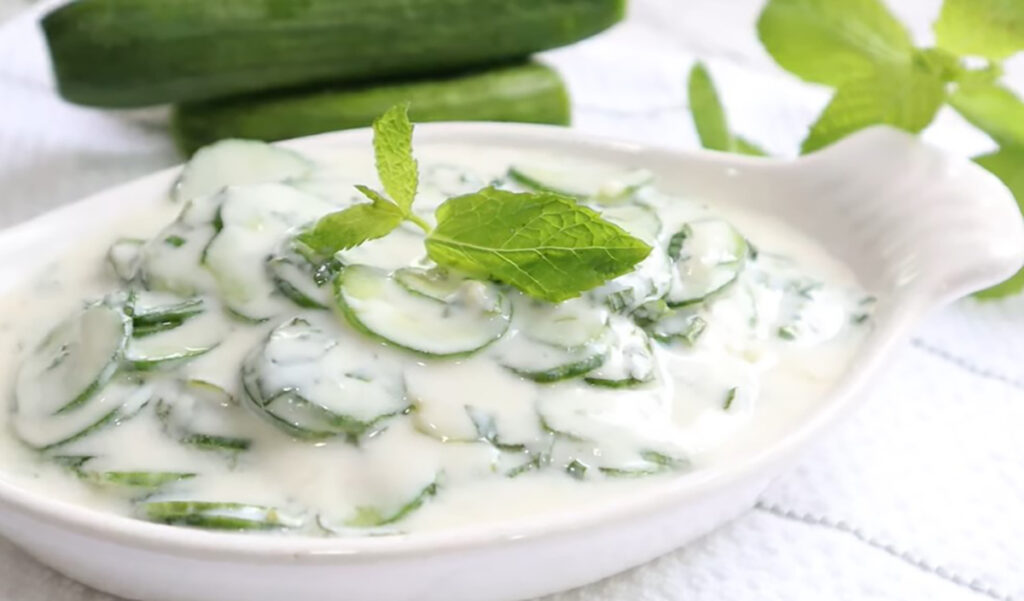 Jajukh | Refreshing Yogurt Salad with Cucumbers and Mint in a white long meza plate with yogurt cucumbers inside plate accented with a fresh mint leave with a pile of cucumbers in backbround laying ontop of a white stripped kitchen towel.