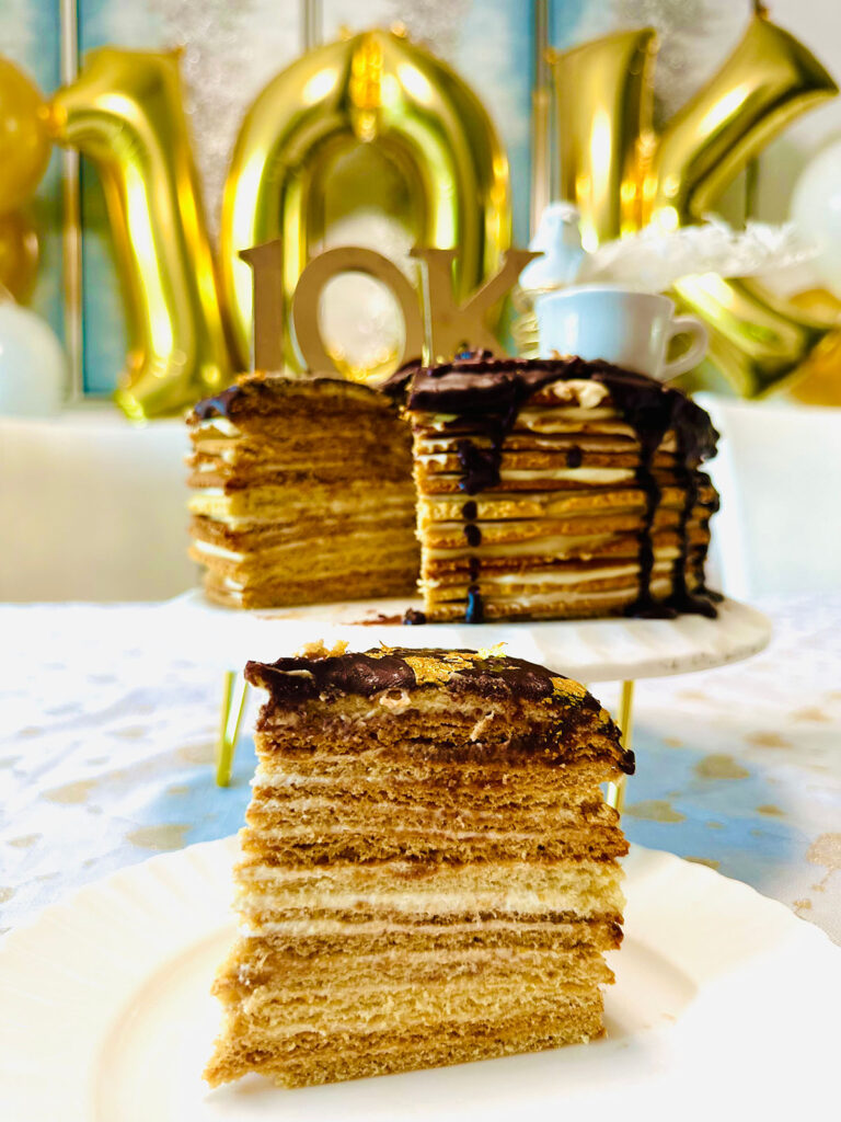 The Classic Birdsmilk Cake (Ptasie Mleczko) sliced cake set infront of the entire cake on a white porcalin cake that has edged ridges around plate with golden numbers of 10k in background on a white tablecloth with golden hearts all over tablecloth.