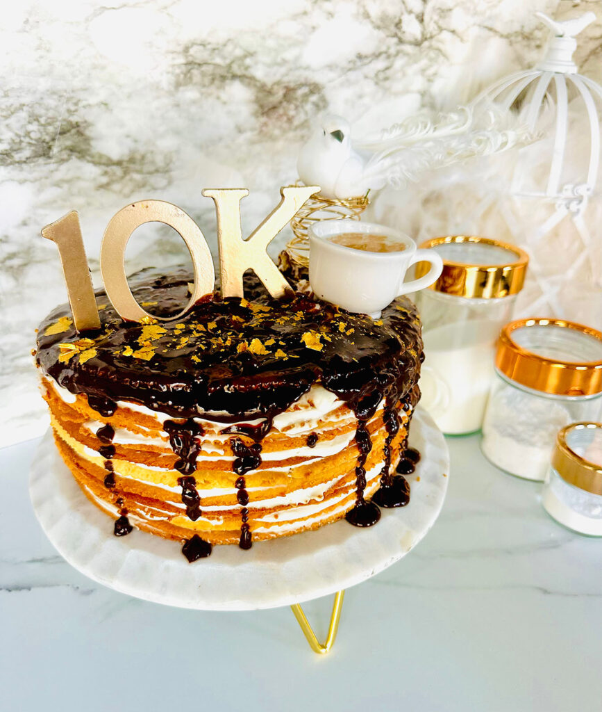 The Classic Birdsmilk Cake (Ptasie Mleczko) layered cake with chocolate dripping sauce over ontop with golden 10k with a white cup and a white bird ontop of the cake with glass with golden lid canisters filled wth sugar, flour, baking soda in them with a white birdcage with white feathers in background on a white countertop.