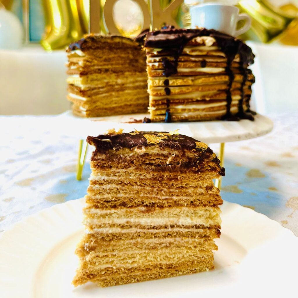 The Classic Birdsmilk Cake (Ptasie Mleczko) sliced cake set infront of the entire cake on a white porcalin cake that has edged ridges around plate with golden numbers of 10k in background on a white tablecloth with golden hearts all over tablecloth.