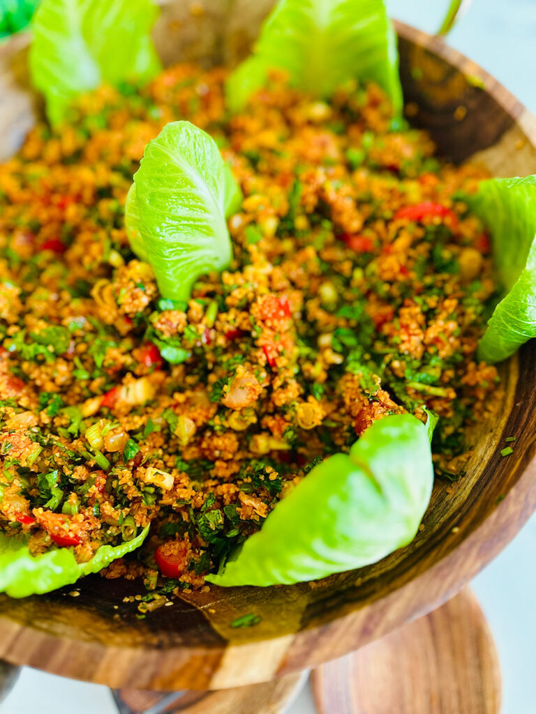 Eech / Itch Bulgur Salad with fresh herbs in a wooden bowl with two wooden spoons laying infront of the bowl with lettuce leaves accenting around bowl with a bowl with green ferns in background as a decor on a white tabletop.