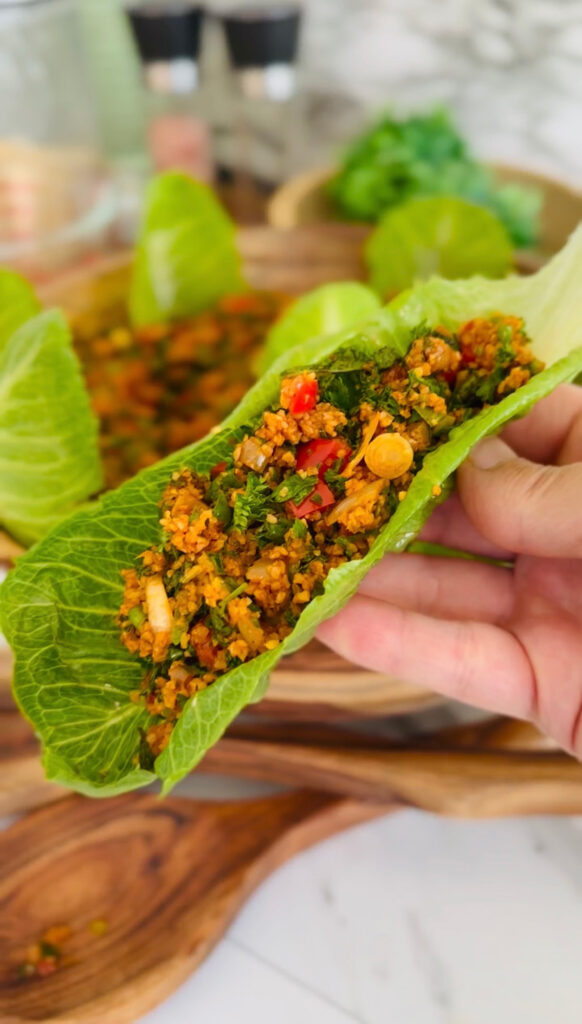 Eech / Itch Bulgur Salad with fresh herbs in a wooden bowl with two wooden spoons laying infront of the bowl with lettuce leaves accenting around bowl with a bowl with green ferns in background as a decor on a white tabletop.