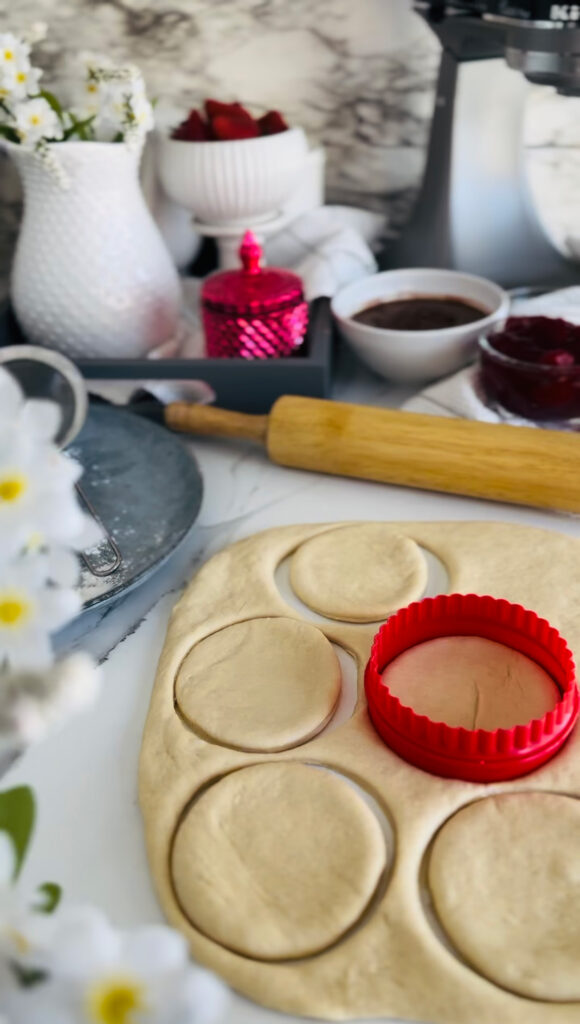 Nutella and Strawberry Filled Donuts (Ponchiks) Dough rolled out and cut with a red round cookie cutter. White with yellow center flowers accenting photo with a blue place with sifter laying ontop. Pink candle with white pitcher and bown in background.
