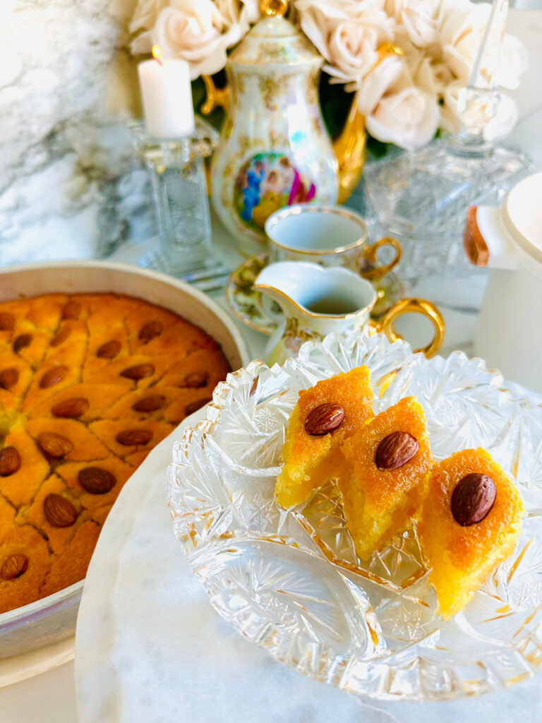 Semolina Cake with Rose Water Syrup - Middle Eastern Basbousa on a crystal platter ontop of a white platter with a silver tin pan basbousa cut into triangled pattern with a vintage tea set in background with a crystal candlestick with a white candle with cream flowers in background