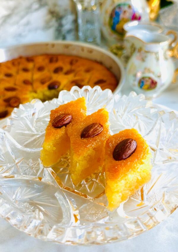 Semolina Cake with Rose Water Syrup - Middle Eastern Basbousa on a crystal platter ontop of a white platter with a silver tin pan basbousa cut into triangled pattern with a vintage tea set in background with a crystal candlestick with a white candle with cream flowers in background