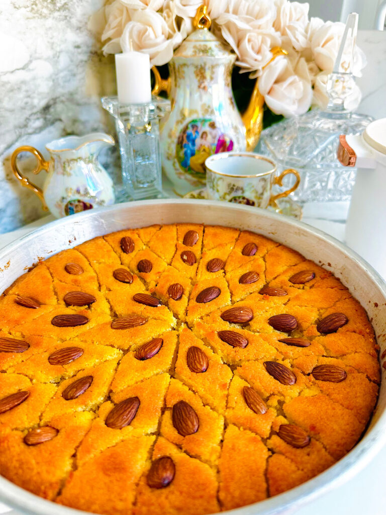 Semolina Cake with Rose Water Syrup - Middle Eastern Basbousa on a crystal platter ontop of a white platter with a silver tin pan basbousa cut into triangled pattern with a vintage tea set in background with a crystal candlestick with a white candle with cream flowers in background
