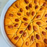 Semolina Cake with Rose Water Syrup - Middle Eastern Basbousa on a crystal platter ontop of a white platter with a silver tin pan basbousa cut into triangled pattern with a vintage tea set in background with a crystal candlestick with a white candle with cream flowers in background