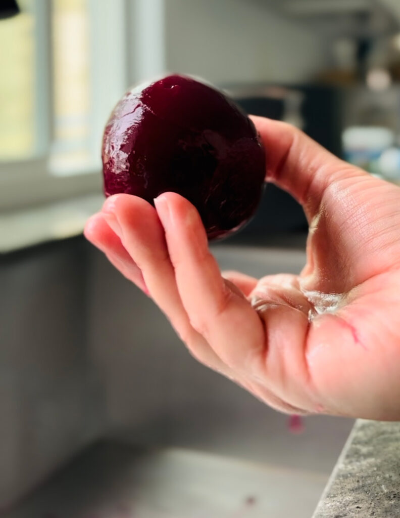Hand holding one peeled beet with a black and stainless steal sink and window in background.