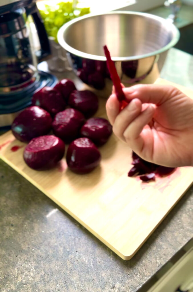 Hand holding a chopped beet strip with beets on a wooden chopping board with a coffee machine and a silver metal mixing bowl in background 