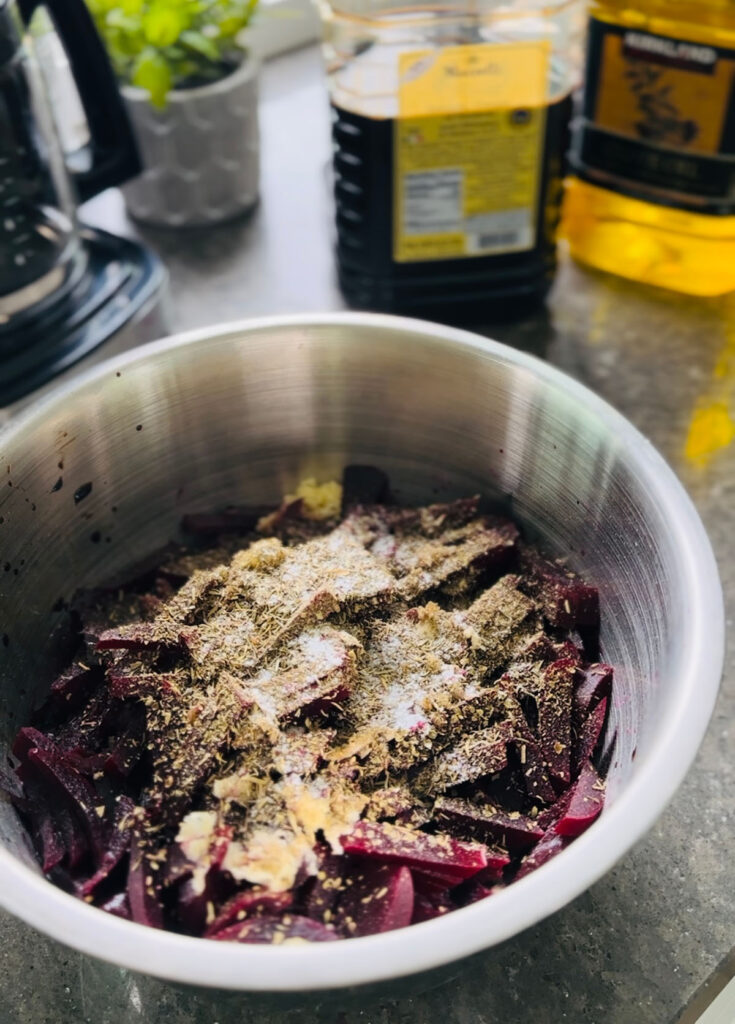 Chopped beets in a metal mixing bowl with seasonings ontop with two bottles in background and coffee machine in background on a black countertop.