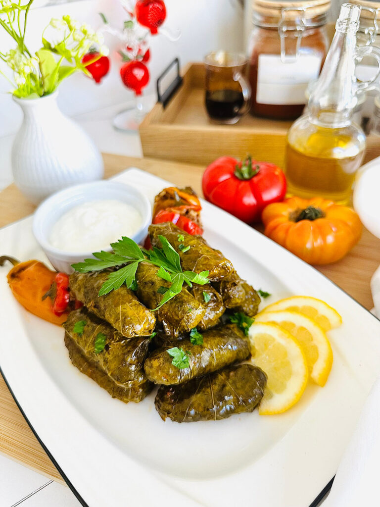 Stuffed Grape Leaves (Dolma / Samra) are displayed on a white platter with black trim around the platter accented with lemon slices, Italian parsley as decor laying ontop of dolma, stuffed bell peppers, and a white round dipping sauce on the platter. Tomatoes wooden tray with a pitcher of pomegranate molasses and a white vase with green flowers in the background with a glass accent decore of pomegranates hanging in the background with an oil container accenting background.