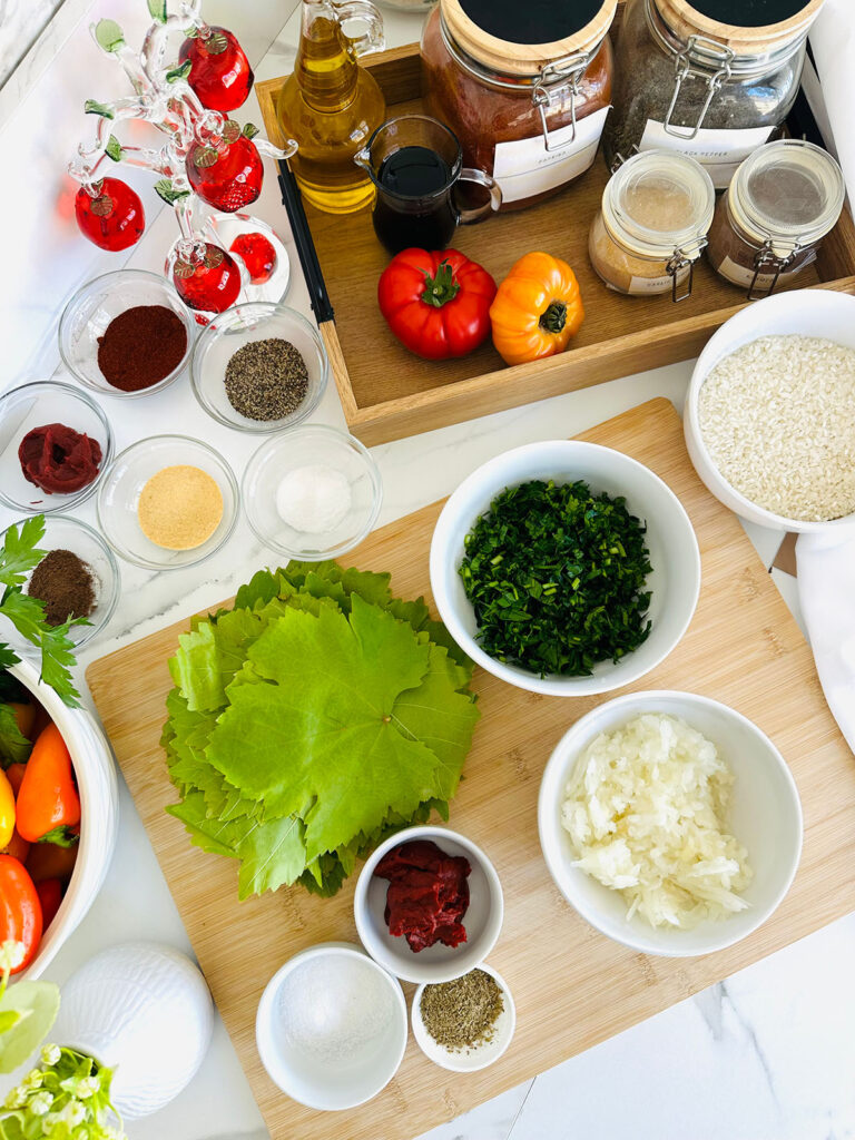 Grape Leaves Dolma / Sarma ingredients on a wooden chopping board in white bowls with tomato, white vase with green flower, glass ornament pomagranet display in background, wooden tray with black handles, canisters, glass oil jar in background.