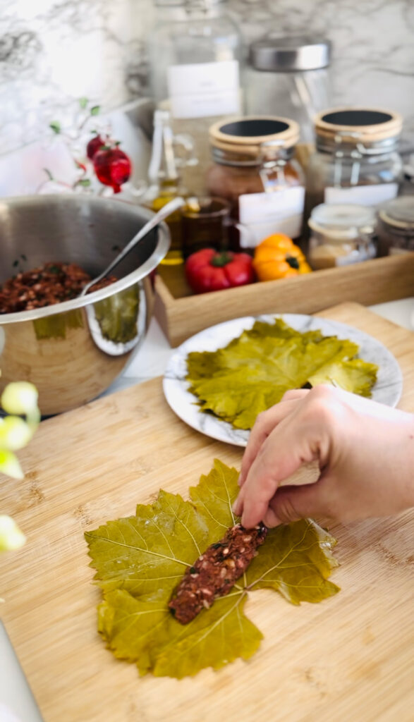 Grape Leaves Dolma / Sarma on a white platter with a black trim piled ontop of each other with stuffed peppers and lemon slices accenting dolma with a white plate with garlic yogurt or sour cream dipping sauce with tomato, white vase with green flower, glass ornament pomegranate display in the background, wooden tray with black handles, canisters, glass oil jar in background.