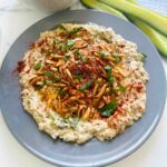 Babaganoush Recipe in a grey platter topped with roasted nuts spice and sliced cucumbers and a white towl on a tabletop.