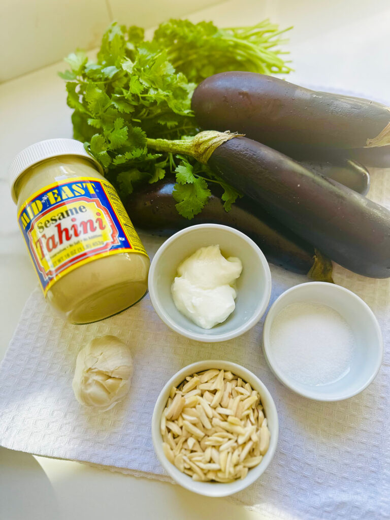 Babaganoush Recipe jar of tahini laying on the counter with 2 eggplants, cilantro with white bowls filled with salt and slivered almonds, yogurt and garlic, all on counter top.