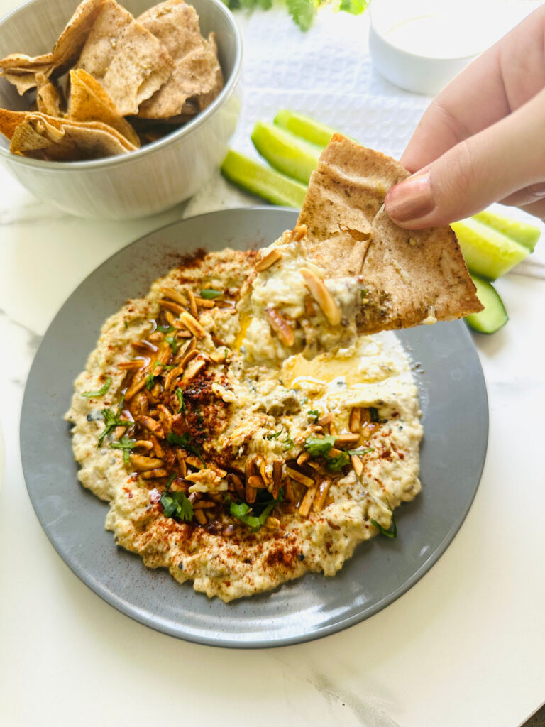 Babaganoush Recipe in a grey platter topped with roasted nuts spices with a bowl of pita chips and sliced cucumbers and a white towl on a tabletop.