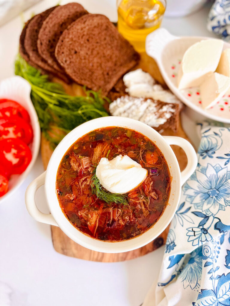 The Best Classic Borscht Soup Recipe white bowl filled with soup filled with borscht with blue and white floral pattern cloth accenting table filled wheat toast, feta cheese on a white plate with red tiny flowers with tomatoes.
