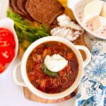 The Best Classic Borscht Soup Recipe white bowl filled with soup filled with borscht with blue and white floral pattern cloth accenting table filled wheat toast, feta cheese on a white plate with red tiny flowers with tomatoes.