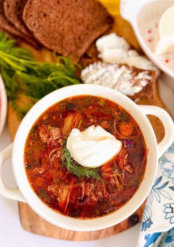 The Best Classic Borscht Soup Recipe white bowl filled with soup filled with borscht with blue and white floral pattern cloth accenting table filled wheat toast, feta cheese on a white plate with red tiny flowers with tomatoes.