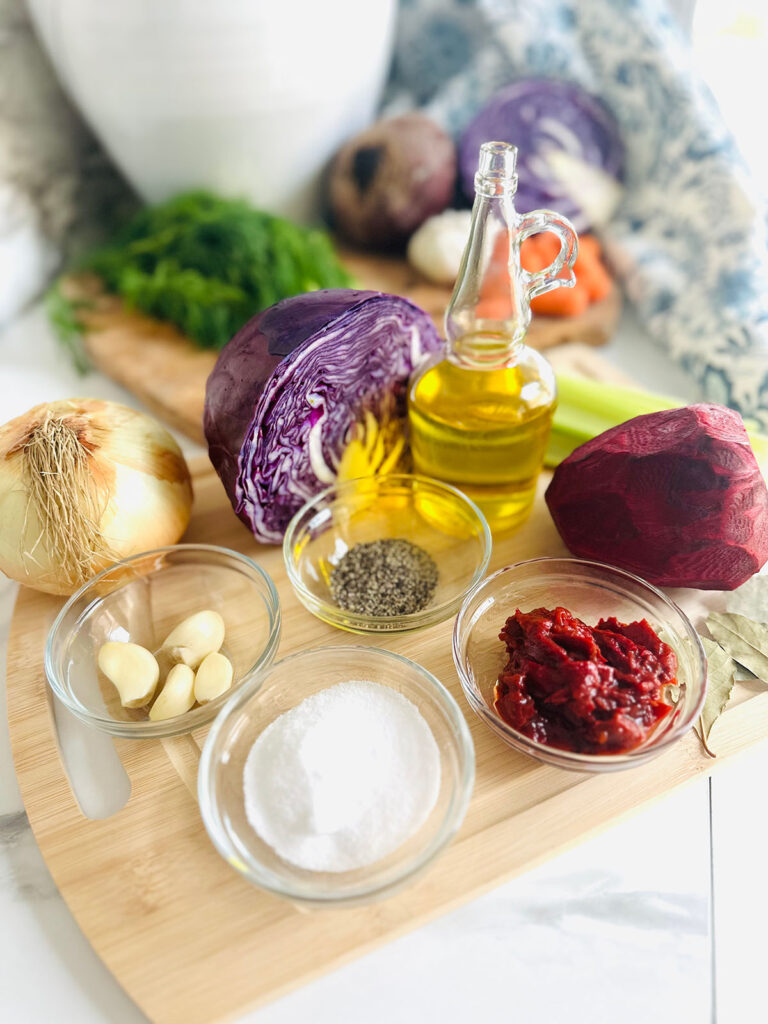 The Best Classic Borscht Soup Ingredients with glass small bowls filled with borsch ingredients on a wooden chopping board fresh vegitables in backgoud with a white vase and blue and white floral fabric in background.