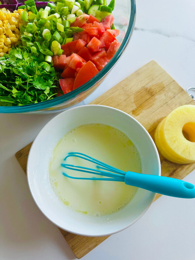Honey Mustard Chicken with Pinapple Salad w/ Orzo Rice bowl white bowl with salad dressing and blue whisk pinapple slices on a wooden chopping board.