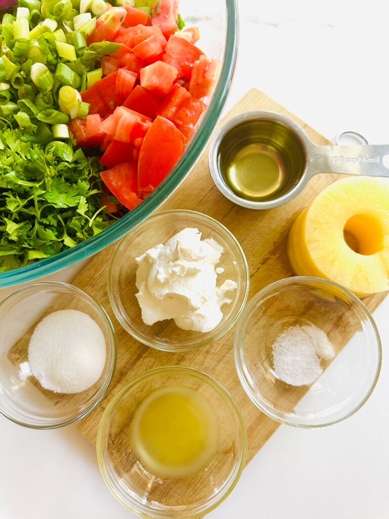 Honey Mustard Chicken with Pinapple Salad w/ Orzo Rice bowl with salad ingredients ontop of a wooden chopping board with a glass bowl filled with veggies, sour cream, pineapple slices, oil in a meacing cup, glass containers of juice, sugar and salt