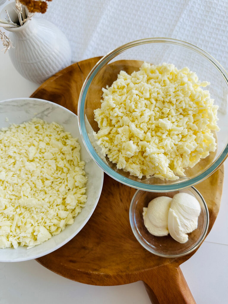 Khachapuri - Georgian Cheese Bread 3 different cheese in white bowl glass bowl on a round wooden board with a white vase in background.