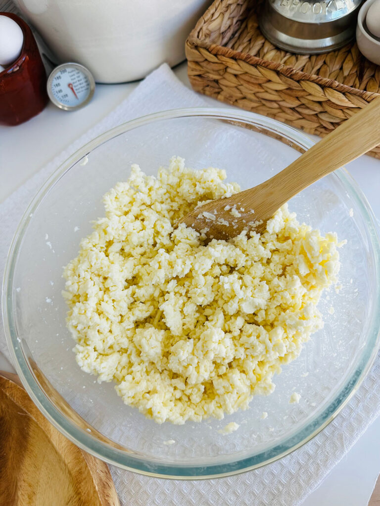 Khachapuri - Georgian Cheese Bread - Flour and yeast mixture in a glass mixing bowl with a white towel underneath with a wooden tray woven basket eggs in a brown bowl flour canister and a thermometer in background