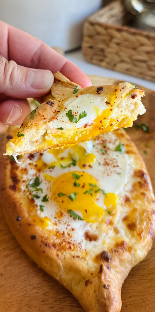 Khachapuri - Georgian Cheese Bread on a round wooden board hand holding a piece of cheese bread that has egg yolk dripping from it with a silver lantern a large glass canister of flour with a wooven basket in background.