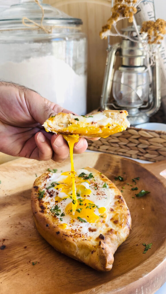 Khachapuri - Georgian Cheese Bread on a round wooden board hand holding a piece of cheese bread that has egg yolk dripping from it with a silver lantern a large glass canister of flour with a wooven basket in background.
