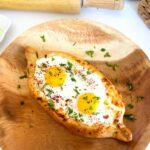 Khachapuri - Georgian Cheese Bread on a round wooden board hand holding a piece of cheese bread that has egg yolk dripping from it with a silver lantern a large glass canister of flour with a wooven basket in background.