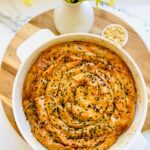 Lamb and Roasted Pine Nut Borek in a white casserole dish ontop of a round wooden board white vase with green flowers with a round plate full of pine nuts on a wooden board on a white counter top.