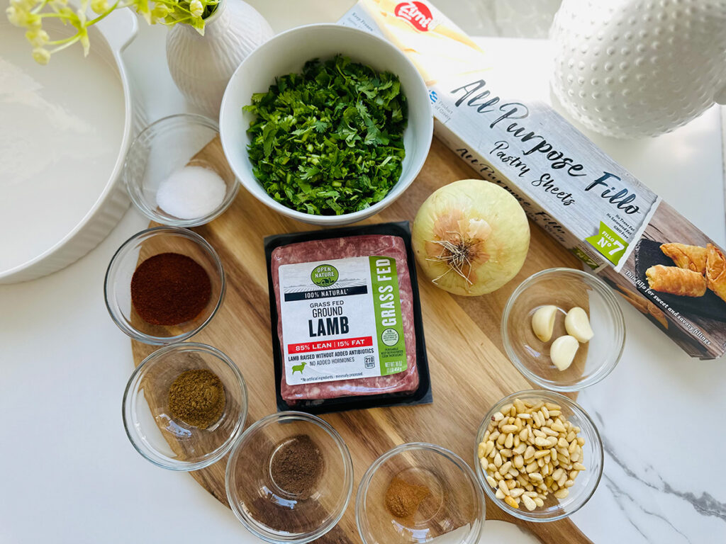 Ingredients for Lamb and Roasted Pine Nut Borek - Small glass containers of spices and white bowls of cilantro a white casserole dish and packaged phyllo dough on top of a round wooden board white vase with green flowers with a round plate full of pine nuts on a wooden board on a white countertop.