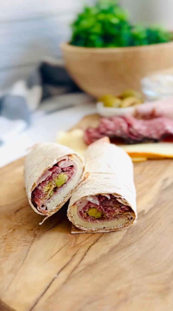 4 Best Wrap Recipes | Perfect Lunch or Dinner Ideas - salami olive lavash wrap on top of a wooden board with salami and cheese in background with a grey and white stripped towel with a wooden bowl