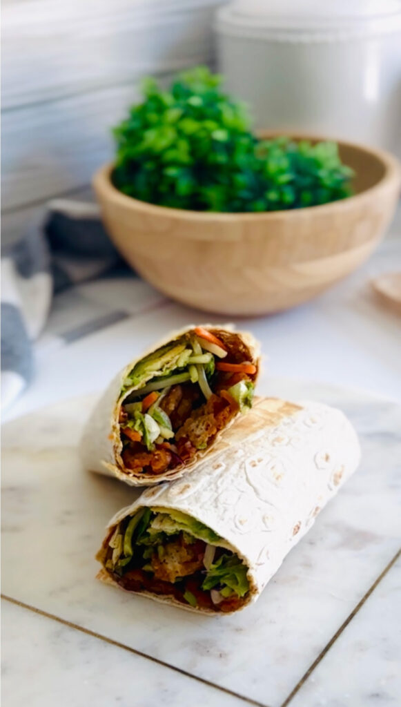 4 Best Wrap Recipes | Perfect Lunch or Dinner Ideas- Teriyaki Chicken wrap on a white marble board with a wooden bowl with green plant, grey and white towel in background.
