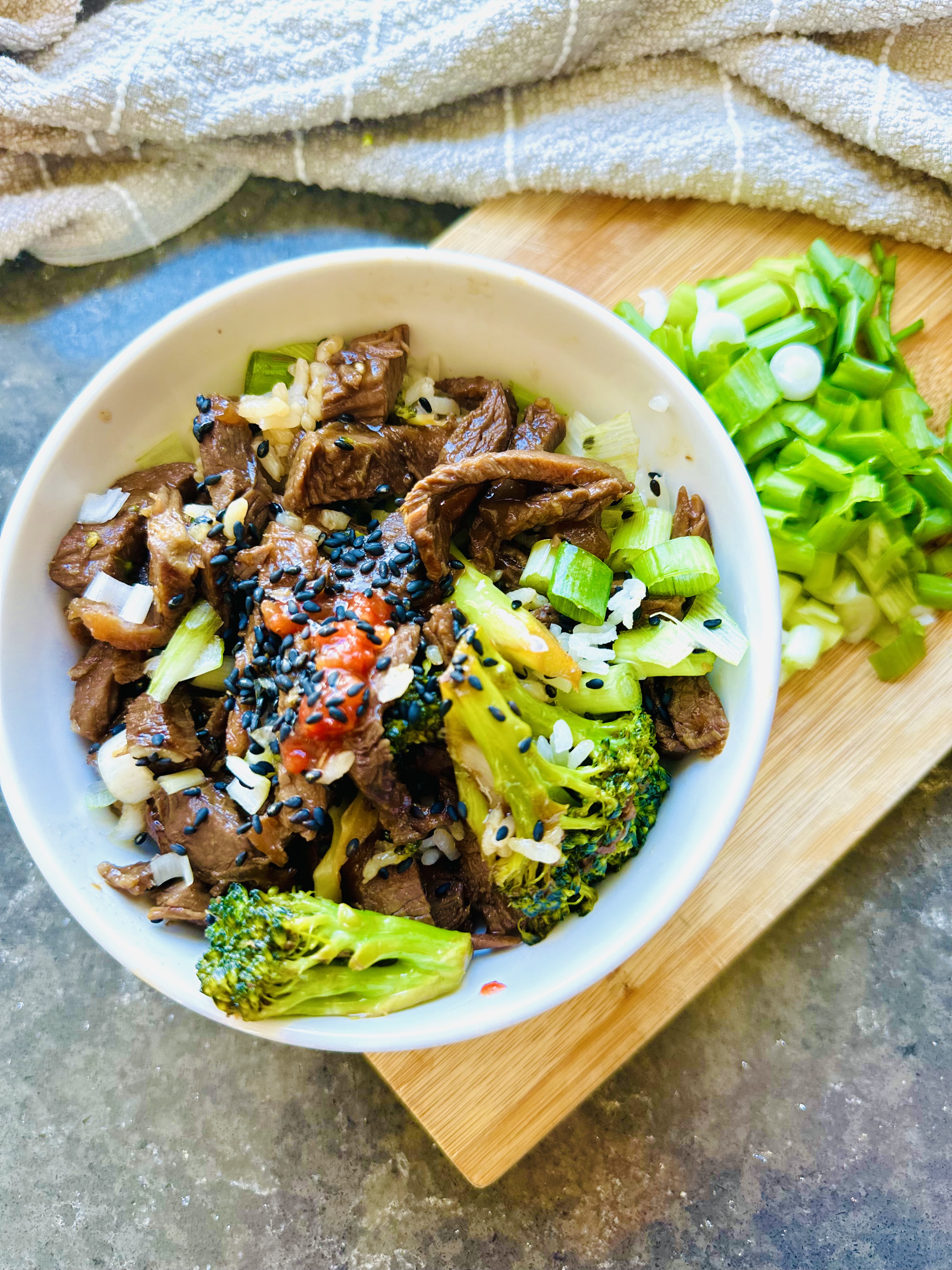 Beef and Broccoli Stir-Fry in a white bowl with beef and broccoli and rice on a wooden chopping board, green onions on board with a grey towl on a black countertop.