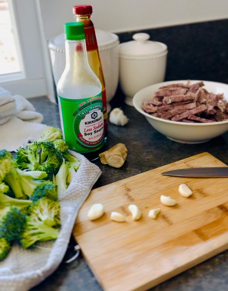 Beef and Broccoli Stir-Fry Wooden chopping board with 6 garlic cloves, white bowl with strips of boiled beef, soy sauce bottle, asian sauce bottle with a towel and some fresh broccoli laying onto with a piece of ginger infront of soy sauce bottle when white canisters in background on a black countertop.