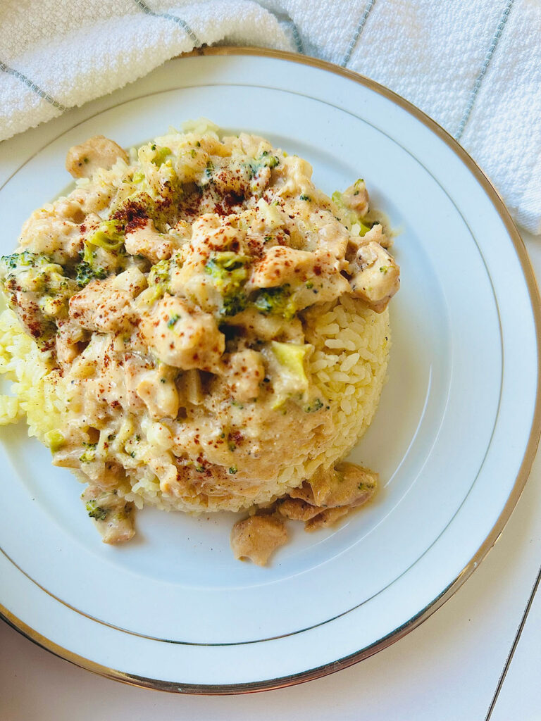Creamy Chicken Broccoli and Rice on a white dinner plate with gold trim and ring around plates. White towel with grey strips accenting plate with a wooden round bowl with broccoli inside bowl with a white pourer on a white table top.