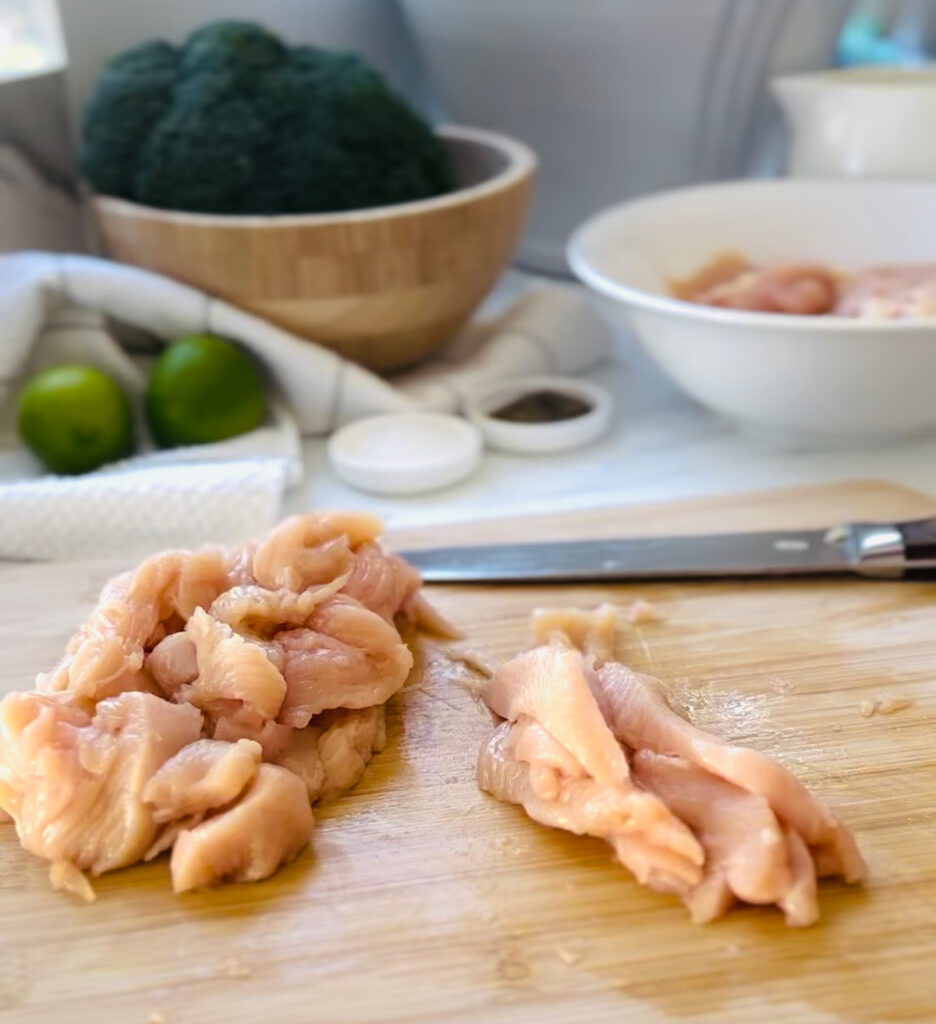 Creamy Chicken Broccoli and Rice wooden chopping board with chopped chicken breast with knife on board. Seasoning saucers, lime on white towel, wooden bowl with broccoli inside, white bowl with chicken in it with a white vase in background on a white countertop.