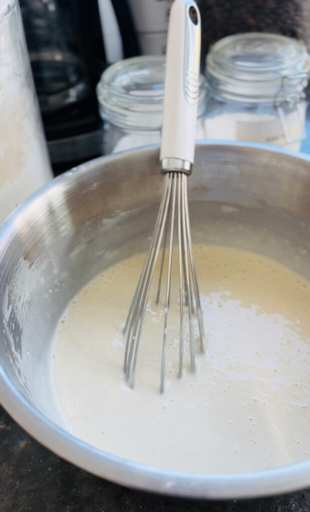 Fluffy Pancake Peach Nut Cluster Toppings - metal mixing bowl with white whisk with pancake batter inside bowl with glass canisters in background.