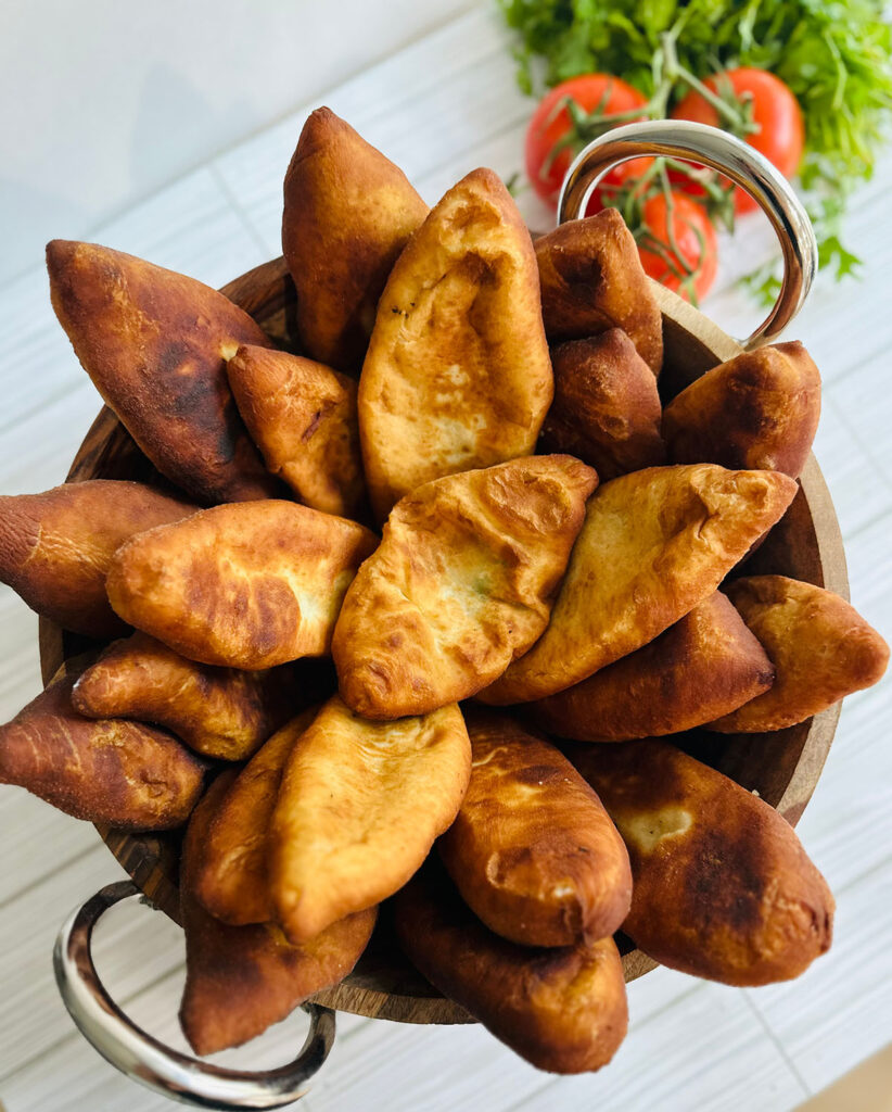 Potato Pirashki / Piroshki wooden bowl with silver handles in fried Pirashki's with fresh tomatoe and cilantro in background.