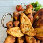 Potato Pirashki / Piroshki wooden bowl with silver handles in fried Pirashki's with fresh tomatoe and cilantro in background.