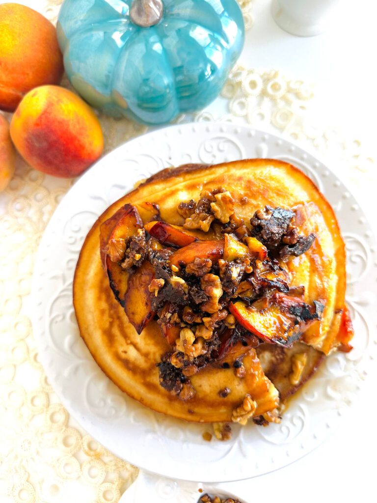 Fluffy Pancake Peach Nut Cluster Toppings on top of a white platter with peache and a turquoise pumpkin in background with white canisters and white pitcher of syrup in background