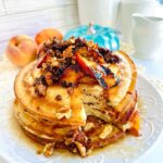 Fluffy Pancake Peach Nut Cluster Toppings on top of a white platter with peache and a turquoise pumpkin in background with white canisters and white pitcher of syrup in background